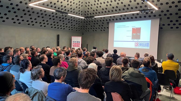 “Multitalent Beton” (“La versatilità del calcestruzzo”): un grande successo! Molte persone assistono a una presentazione in una stanza moderna con soffitto forato.