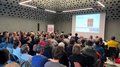 “Multitalent Beton” (“La versatilità del calcestruzzo”): un grande successo! Molte persone assistono a una presentazione in una stanza moderna con soffitto forato.