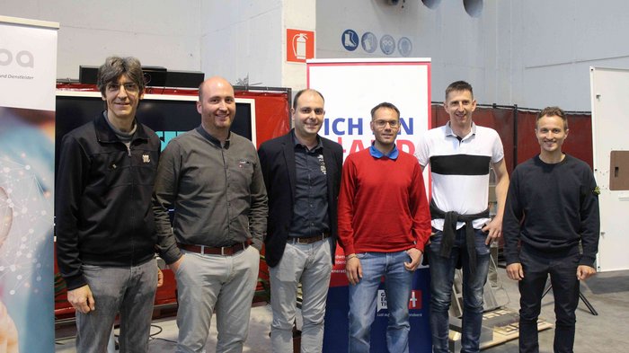 Eletto il nuovo presidente della sezione di mestieri Sei uomini in piedi in fila in un capannone davanti a striscioni pubblicitari