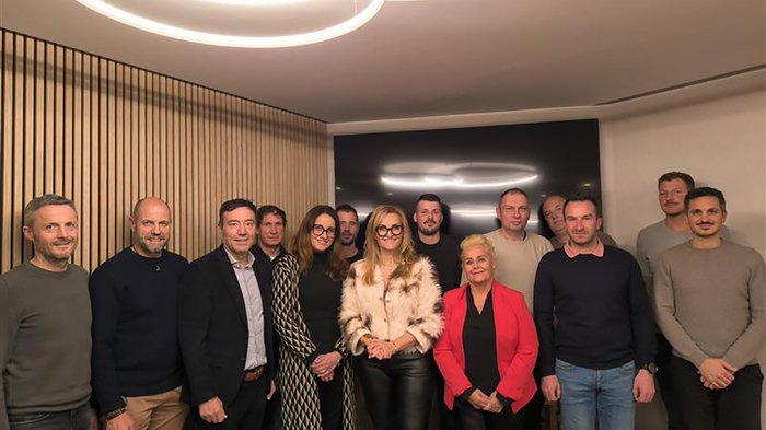 Austausch, Kontinuität und Blick nach vorn Gruppenfoto von 14 Personen in einem modernen Raum mit Holzvertäfelung und Deckenleuchten