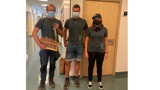 News Drei Personen mit Masken stehen im Flur, eine hält ein Holzspielzeug