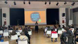 News Frau hält Vortrag vor sitzenden Schülern in einem Saal mit Präsentation