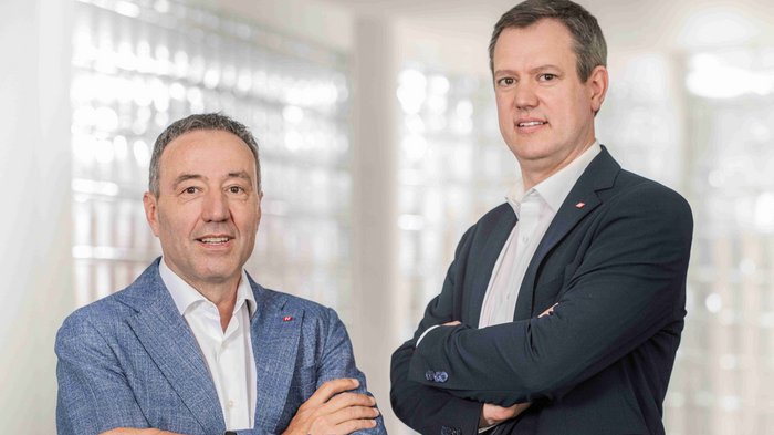 Neuerungen im öffentlichen Auftragswesen: Chancen für Südtirols Handwerksbetriebe Zwei Männer in Anzügen mit verschränkten Armen stehen vor hellem Hintergrund