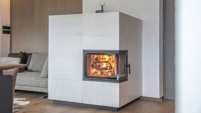 Inspiration, Austausch und neue Impulse beim Textil-Event 2025 Weißer Kaminofen mit loderndem Feuer neben einem grauen Sofa im Wohnzimmer
