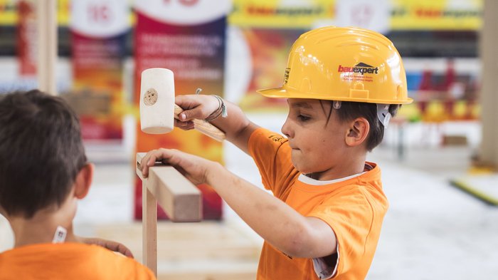 "Avventura artigianato" entusiasma a Merano: i giovani talenti vivono l’artigianato da vicino Bambino con casco giallo che batte il legno in un cantiere