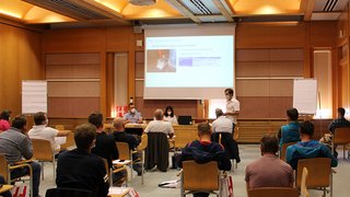 News Menschen sitzen mit Abstand in einem Seminarraum und hören einem Vortrag zu.