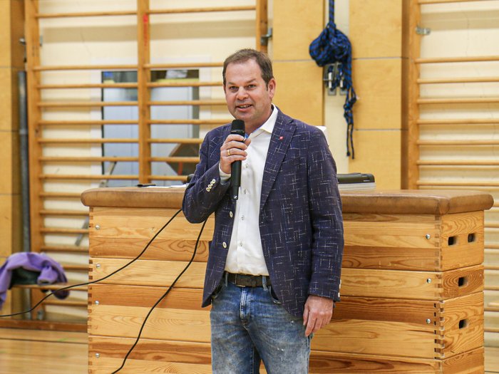 Kreative Spielideen: Preisverleihung für "Clever gebaut" Mann spricht mit Mikrofon in einer Turnhalle vor Holzmöbeln