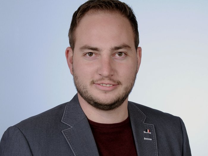 Investitionen in Infrastrukturen Porträt eines lächelnden Mannes mit Bart, grauem Sakko und rotem Hemd vor neutralem Hintergrund