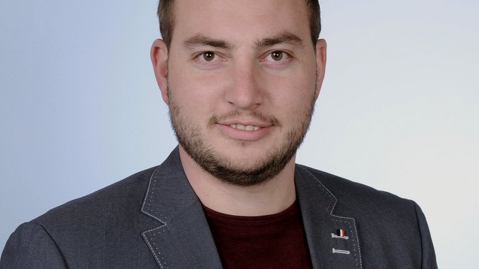 Tiefbauunternehmer: Preisanstiege als größte Herausforderung Porträt eines lächelnden Mannes mit Bart, grauem Sakko und rotem Hemd vor neutralem Hintergrund