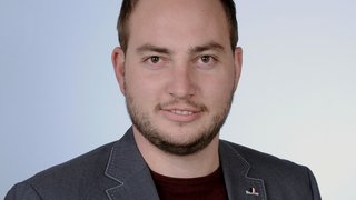 News Porträt eines lächelnden Mannes mit Bart, grauem Sakko und rotem Hemd vor neutralem Hintergrund