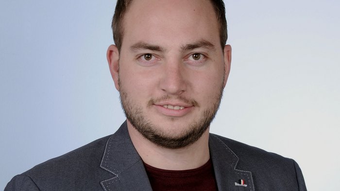 Investitionen in Infrastrukturen Porträt eines lächelnden Mannes mit Bart, grauem Sakko und rotem Hemd vor neutralem Hintergrund