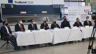 News Sieben Männer mit Masken sitzen an einem langen Tisch vor Kamera in Fabrikumgebung