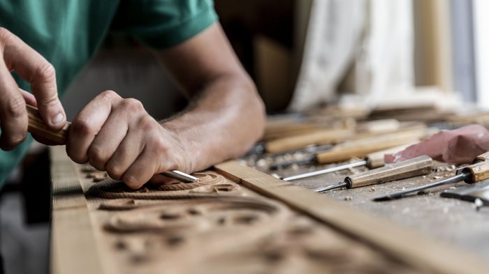 © Alan Bianchi Handwerker schnitzt Holzverzierung mit Meißel am Arbeitstisch