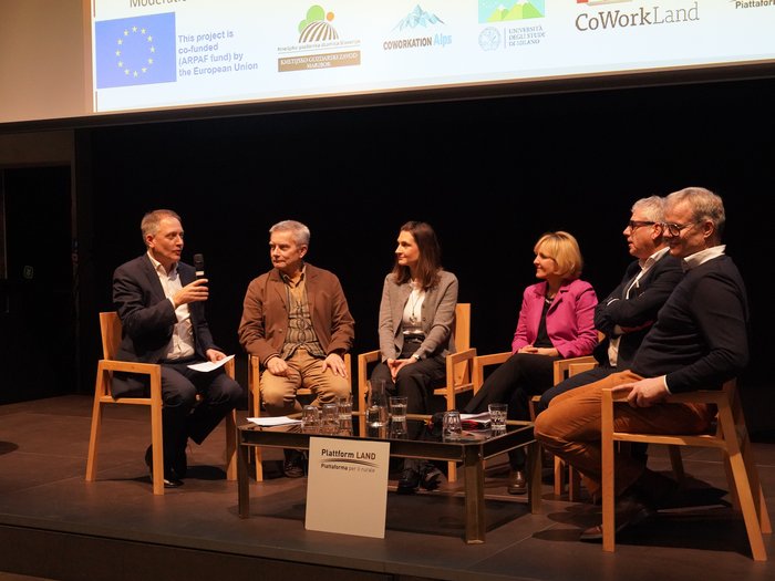 Crescente domanda di coworking e assistenza all’infanzia Discussione in panel con sei persone sedute su palco, tavolo con bicchieri davanti