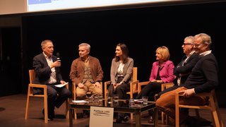 News Podiumsdiskussion mit sechs Personen bei einer Veranstaltung, Tisch mit Gläsern im Vordergrund