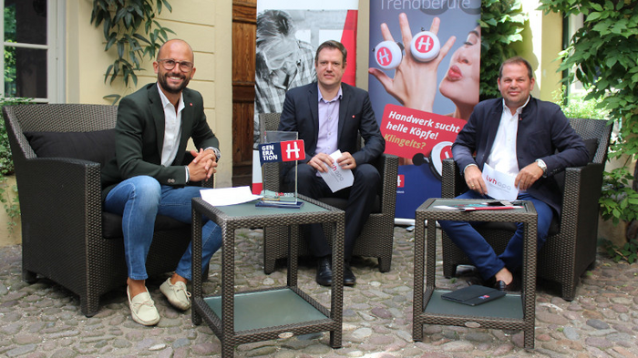 Resilienz und Adaptionsfähigkeit sind Stärken des Handwerks Drei Männer sitzen auf Rattanmöbeln vor Bannern bei einer Außenveranstaltung