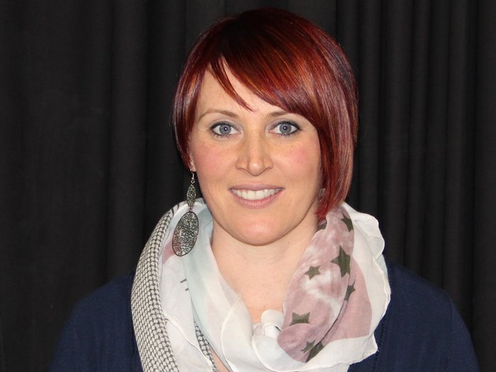 „Es wird Zeit, dass wir wieder aufsperren dürfen!“ Frau mit rotem Kurzhaarschnitt und gemustertem Schal vor schwarzem Hintergrund