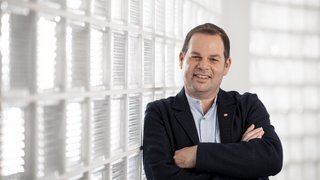 News Lächelnder Mann lehnt an Glasmauer mit verschränkten Armen in hellem Raum