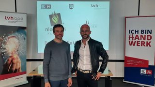 News Zwei Männer stehen vor einem Bildschirm und Bannern bei einer Handwerksveranstaltung.