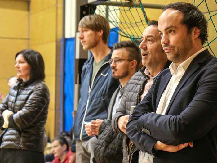 Kreative Spielideen: Preisverleihung für "Clever gebaut" Fünf Personen beobachten konzentriert ein Sportereignis in der Halle