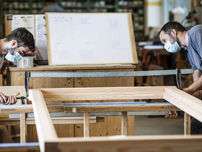 „Nie zuvor wurden wir auf so eine harte Probe gestellt“ Zwei Handwerker mit Masken montieren einen Holzrahmen in einer Werkstatt