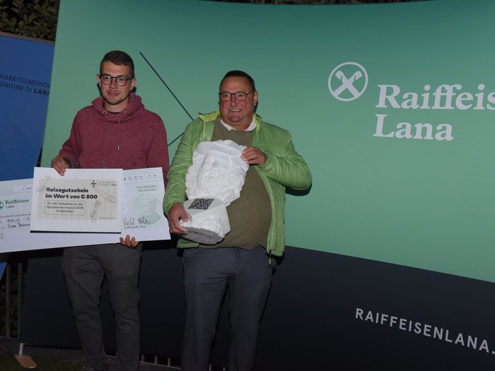 Ein starkes Zeichen für das junge Handwerk – Erfolgreiche Premiere des 1. Förderpreises Junges Handwerk in Lana Zwei Männer halten Preis und Statue vor Raiffeisen Lana Werbewand