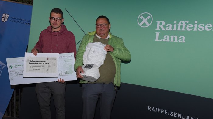 Ein starkes Zeichen für das junge Handwerk – Erfolgreiche Premiere des 1. Förderpreises Junges Handwerk in Lana Zwei Männer halten Preis und Statue vor Raiffeisen Lana Werbewand