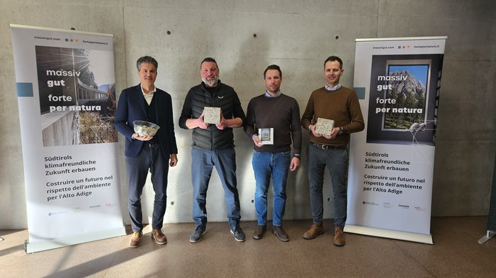Presentata l‘iniziativa “forte per natura”: per un metodo costruttivo rispettoso del clima ed efficiente nella gestione delle risorse Quattro uomini mostrano materiali da costruzione ecologici davanti a una parete di cemento