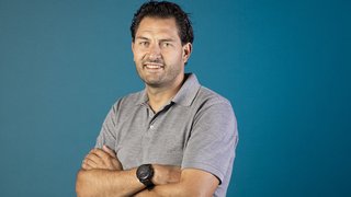 News Mann mit verschränkten Armen und grauem Poloshirt vor blauem Hintergrund