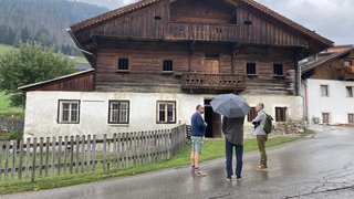 News Drei Männer stehen vor einem alten Holzhaus bei regnerischem Wetter.