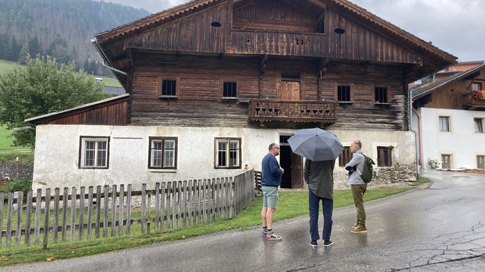 Plattform Land setzt neue Akzente für 2025 Drei Männer stehen vor einem alten Holzhaus bei regnerischem Wetter.