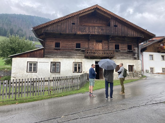 Plattform Land setzt neue Akzente für 2025 Drei Männer stehen vor einem alten Holzhaus bei regnerischem Wetter.