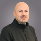 Andreas Acler Ritratto di un uomo sorridente con testa calva e giacca nera su sfondo grigio