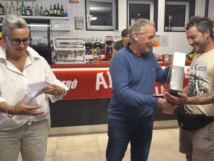 Gemeinsam unterwegs, gemeinsam stärker: Was ein guter Dachdecker-Tag braucht Ein älterer Mann überreicht einem Mann einen Preis in einer Bar, während eine Frau Unterlagen hält
