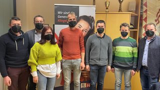News Sieben Personen mit schwarzen Masken stehen in einem Raum vor einem Banner und einem Schrank mit Pokalen