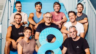 News Gruppe von neun Personen sitzt auf blauen Treppen, hält große blaue Zahl 8