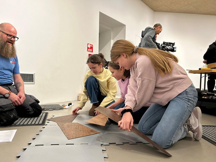 40 Betriebe, 100 Jugendliche, 15 Berufe Drei Mädchen legen Holzpaneele auf weiche Bodenmatten im Raum