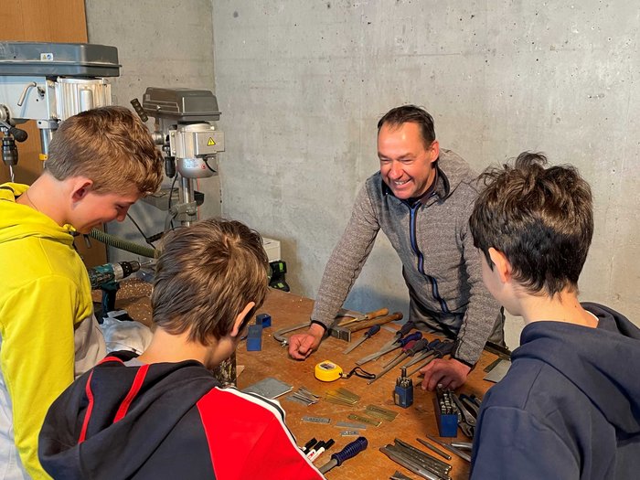 Praktische Berufe kennenlernen – Sculeis dla scoloa mesana mpera a cunescer l'artijanat Erwachsener erklärt Jugendlichen Werkzeuge auf Werkbank in Werkstatt
