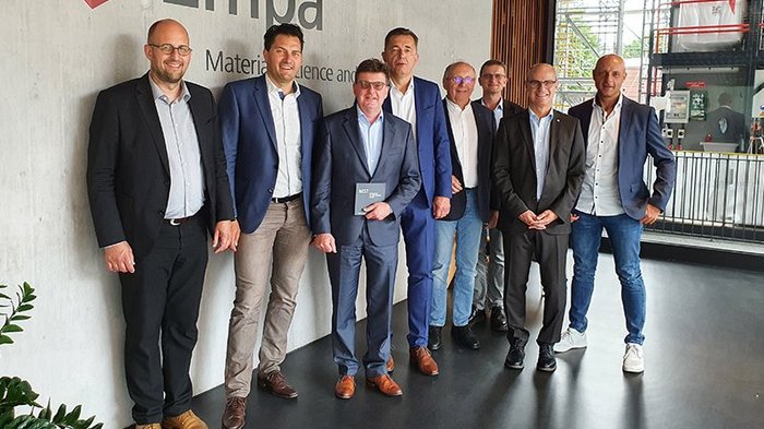 Fachexpertentreffen in Zürich Acht Männer in Anzügen stehen in einem modernen Innenraum an einer Wand