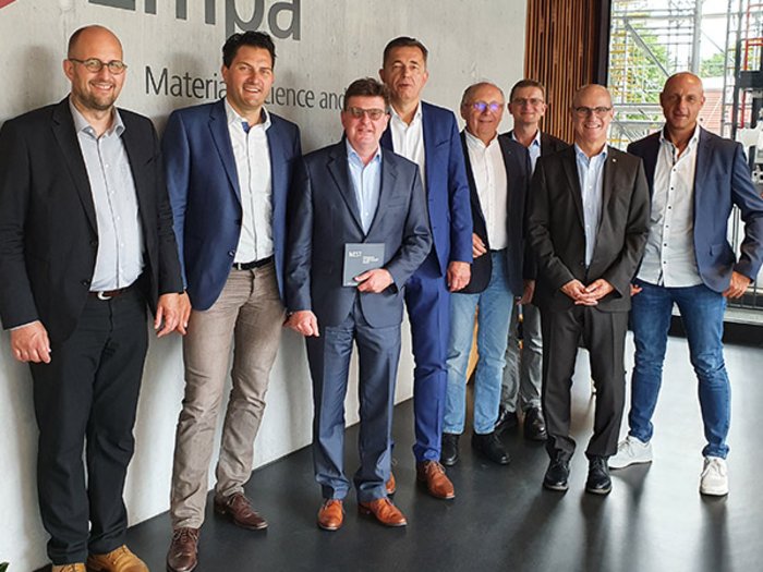 Fachexpertentreffen in Zürich Acht Männer in Anzügen stehen in einem modernen Innenraum an einer Wand