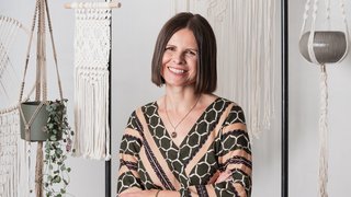 News Frau sitzt lächelnd vor Makramee-Wandbehängen und Pflanzenhalterungen