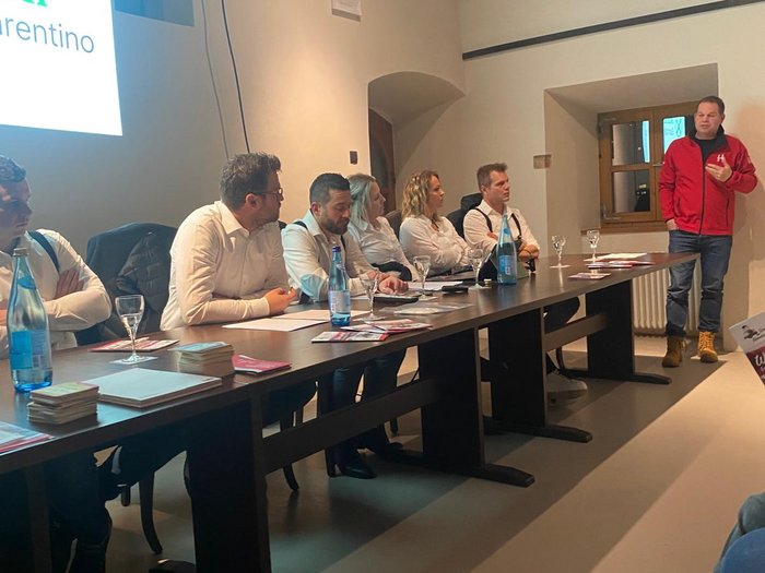 Gute Bilanz der Sarner Handwerker/innen Personen sitzen an einem Tisch und hören einem stehend Vortragenden in rotem Jacke zu