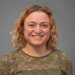 Das lvh-Team Fröhliche Frau mit lockigem blonden Haar und grüner Spitzenbluse vor grauem Hintergrund