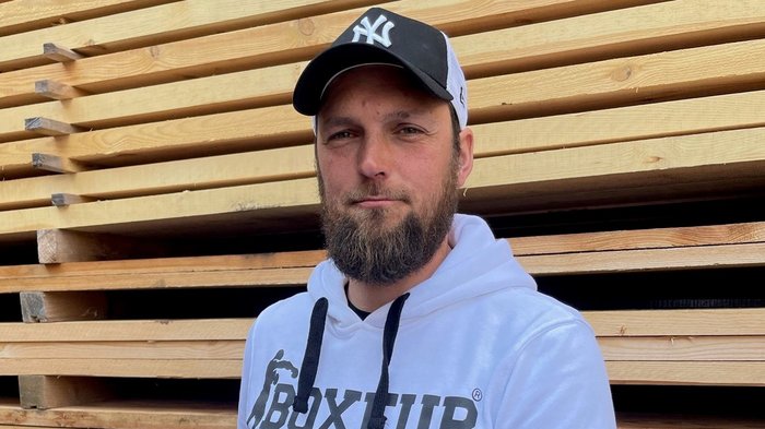 Der Südtiroler Zimmerer-Meistertitel wird jetzt in Deutschland anerkannt Mann mit Baseballkappe und weißem Hoodie hält Dokument vor Holzstapel