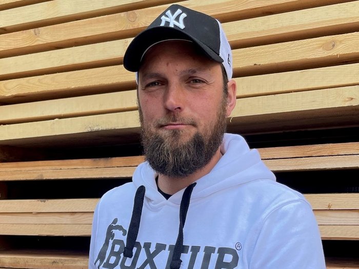 Der Südtiroler Zimmerer-Meistertitel wird jetzt in Deutschland anerkannt Mann mit Baseballkappe und weißem Hoodie hält Dokument vor Holzstapel