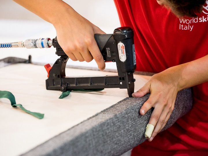 Tapezierer - Raumausstatter Person befestigt grauen Stoff mit einem Tacker an einem Möbelstück