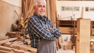 News Älterer Handwerker in Schürze in einer Holzwerkstatt mit Holzplatten
