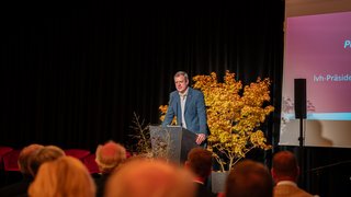 News Mann hält Rede an Podium vor Publikum mit gelbem Baum im Hintergrund