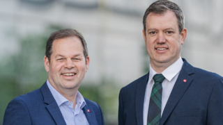 News Zwei Männer in Anzügen lächeln vor einem unscharfen Hintergrund