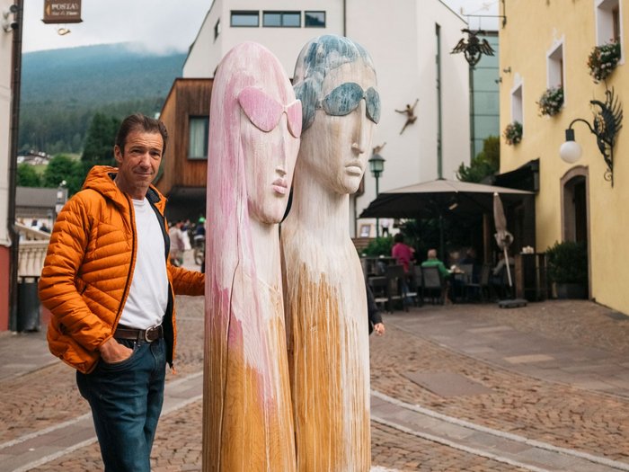 IDEA UNIKA 2025: 6ª edizione della Biennale d’arte nei centri dei paesi della Val Gardena Uomo con giacca arancione accanto a grandi sculture di legno con occhiali da sole in strada pedonale